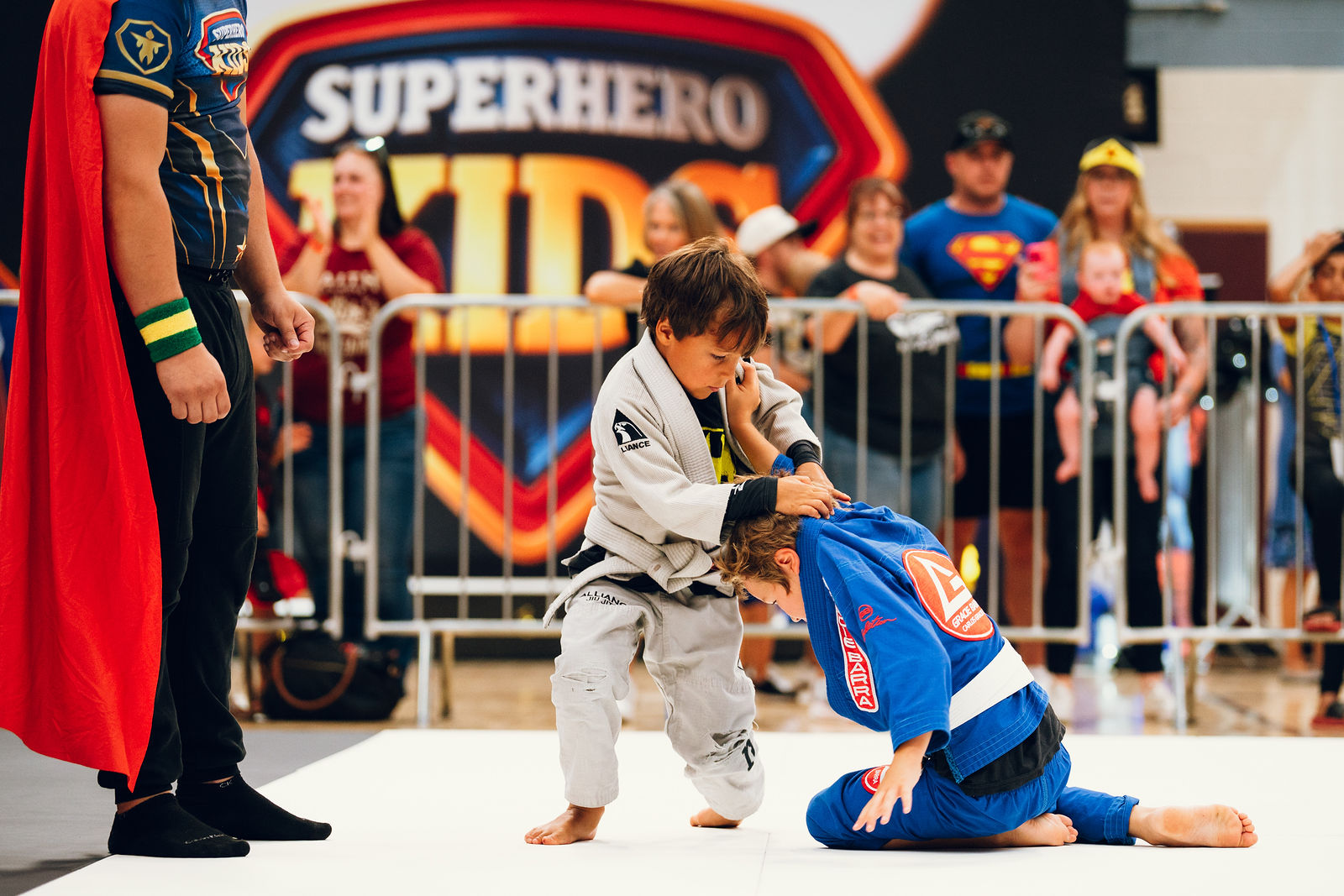 A coach supporting a young athlete from the corner of the mat